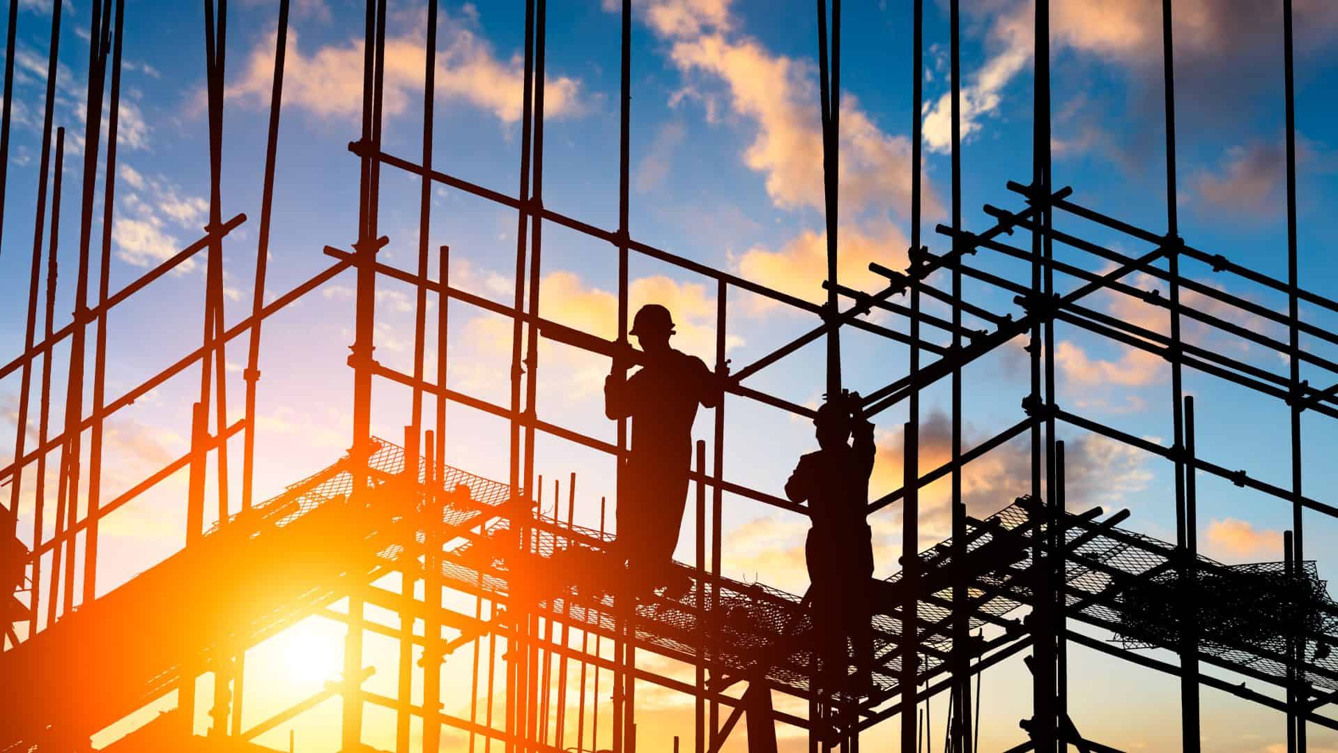 Construction workers stand on scaffolding in front of a setting sun.